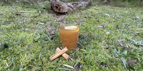 A hot toddy made with black tea and spiced rum sitting on a grass flat with a log in the background.